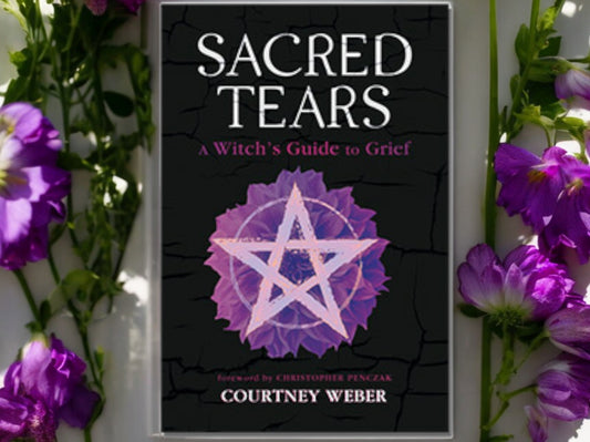 A picture of Sacred Tears: A Witch's Guide to Grief on a white surface with purple flowers.